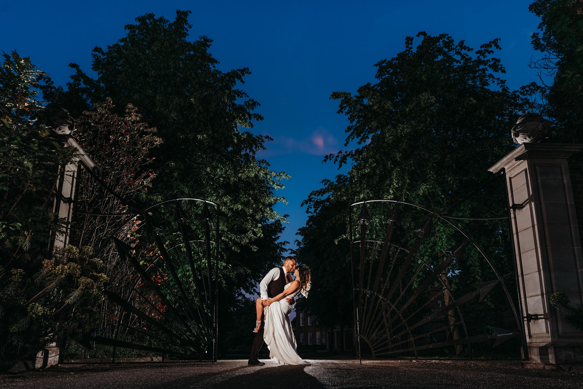 Wedding photograph of groom dipping the bride at the front gates outside Colwick Hall in Nottingham. Beautifully lit with a flash at the front so the sky is blue and the hall behind them.