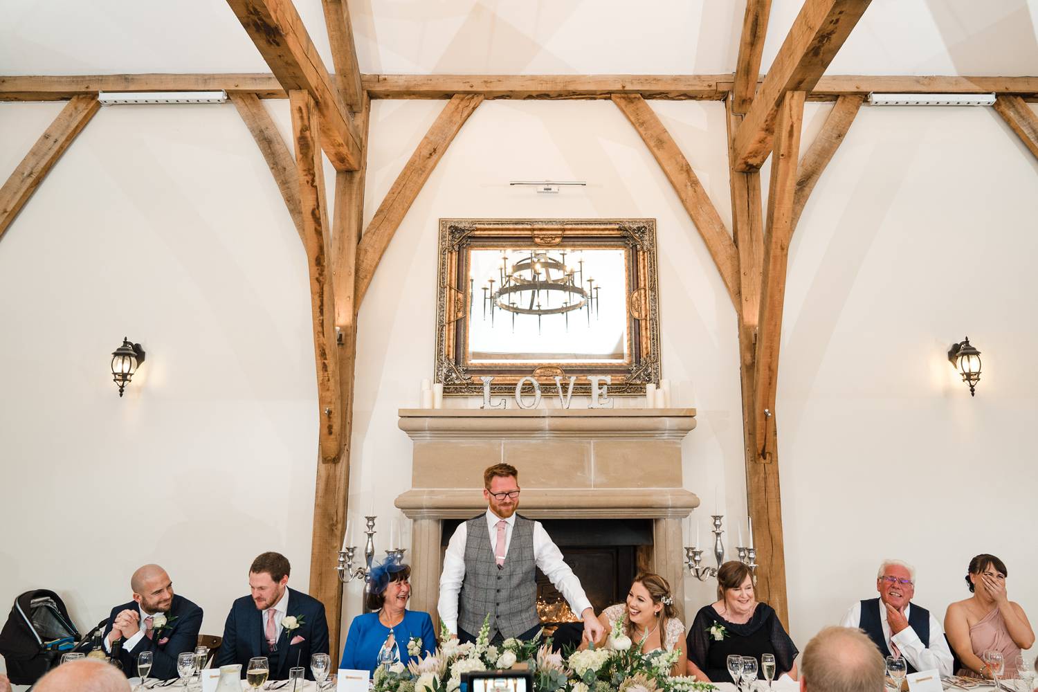 A wide photo showing all the wedding party sat at the top table at Swancar Farm during the groom's speech. The bride is laughing whilst the groom puts his hand on her shoulder. The maid of honour covers her mouth with laughter