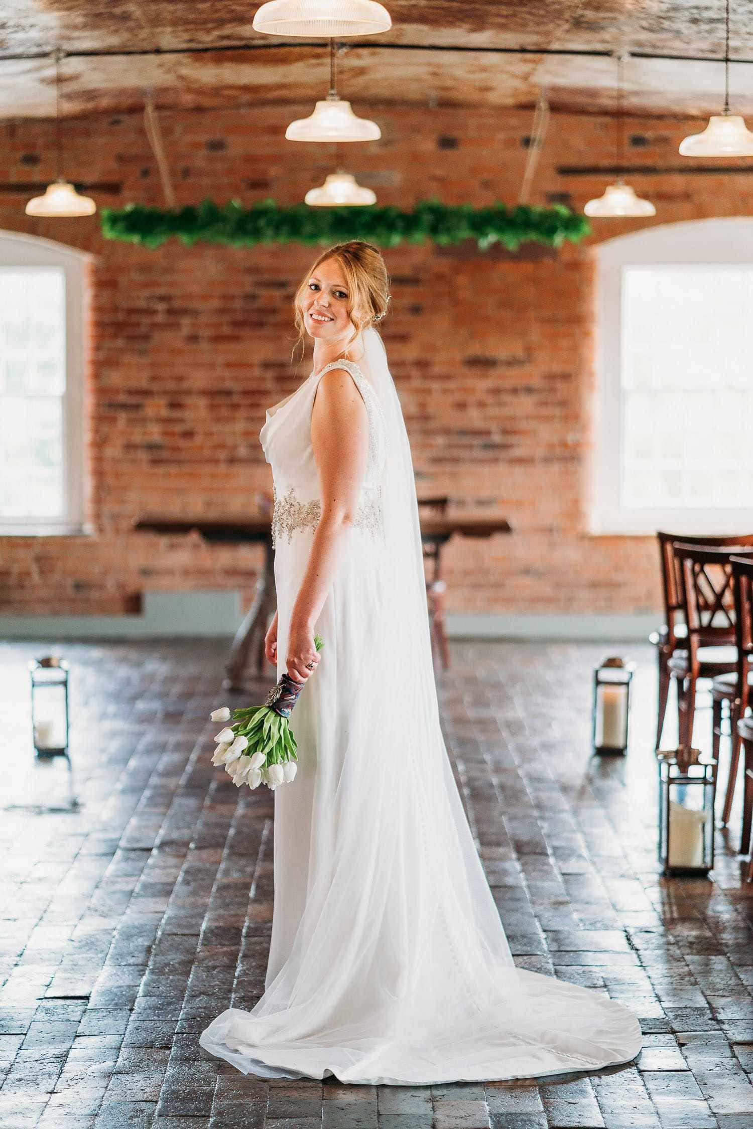 beautiful bride stands in the ceremony room at the west mill holding her bouquet and smiling at the camera