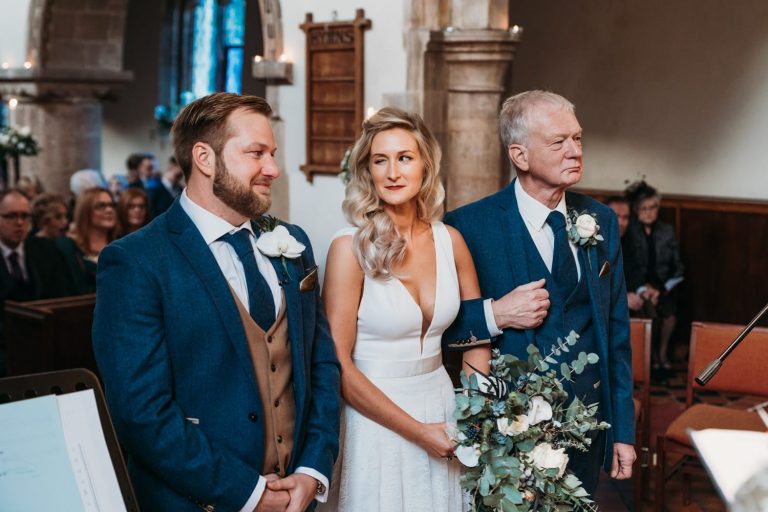 Bride winks at the groom as she walks down the aisle with dad at church