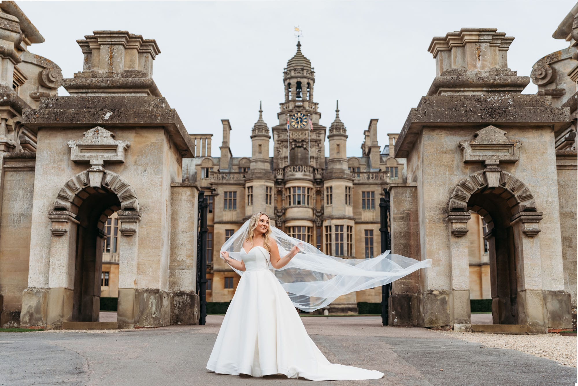 Bride holding her veil blowing in the wind whilst stood at the gates of Harlaxton Manor