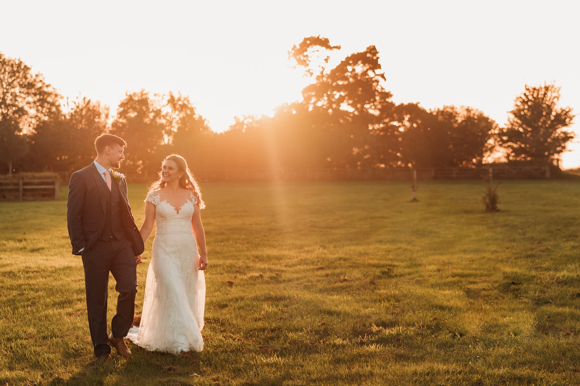 Bride in white dress walking in the field at Mythe Barn with her groom in blue suit with golden hour sunshine behind them