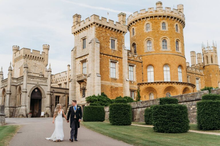 Bride and groom walking hand in hand with Belvoir Castle behind them after their wedding