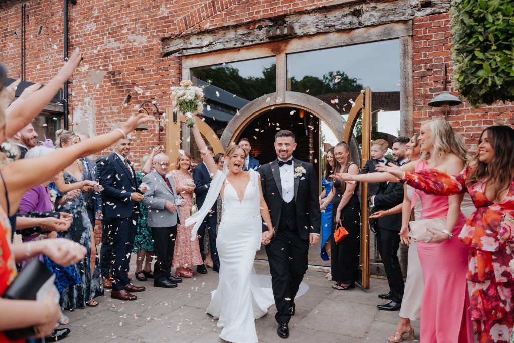Couple cheer and bride holds bouquet in the air whilst they walk through a confetti tunnel at Hazel Gap Barn