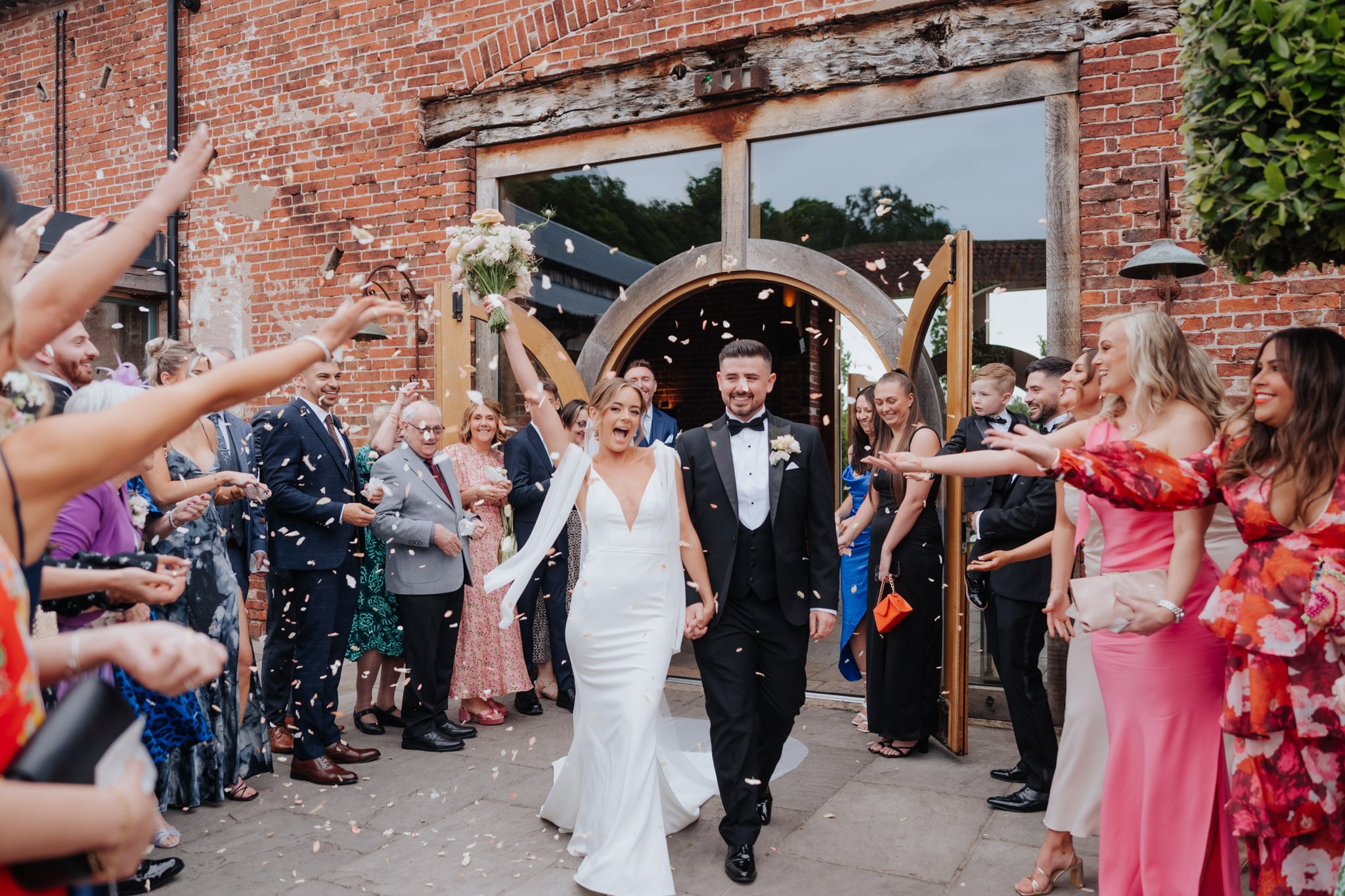 Couple cheer and bride holds bouquet in the air whilst they walk through a confetti tunnel at Hazel Gap Barn