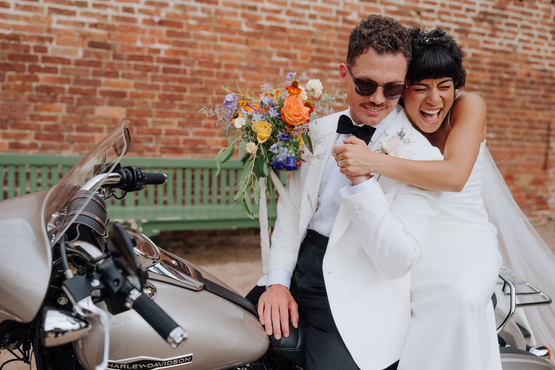 A joyful bride in a white dress hugs and laughs with her groom, who is wearing a white tuxedo jacket and sunglasses, as they sit together on a Harley-Davidson motorcycle. Captured by a Nottingham Wedding Photographer, the bride holds a colorful bouquet.