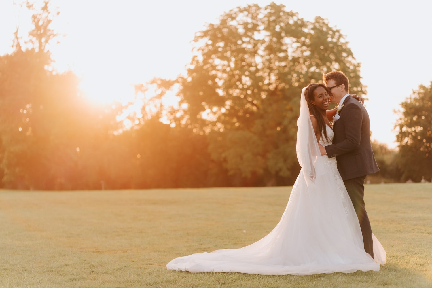 Bride in white dress laughs as groom tells her a joke during golden hour photography session at Stubton Hall