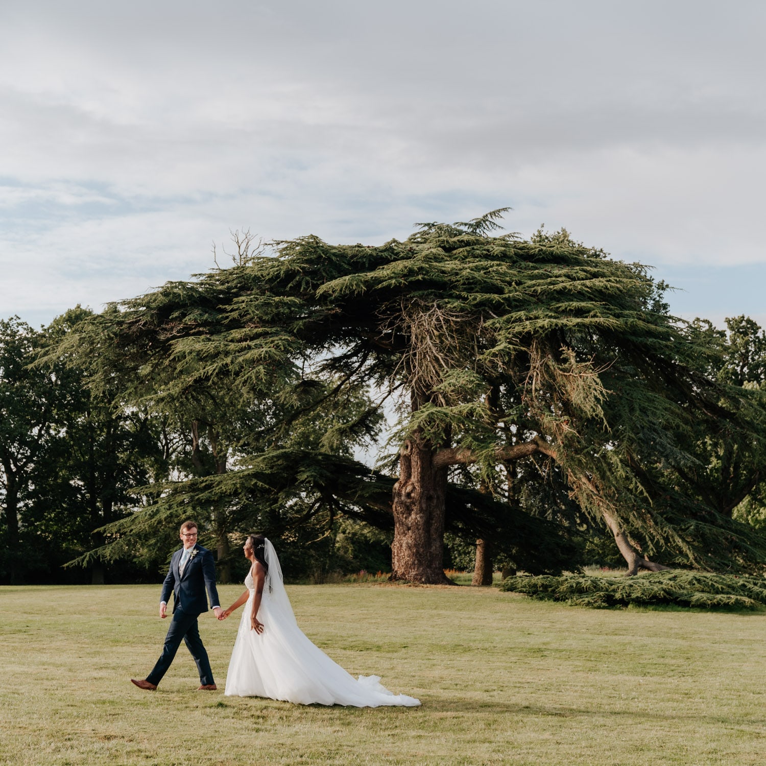 Groom in a blue suit leading the bride on a walk past the cedar tree outside Stubton Hall
