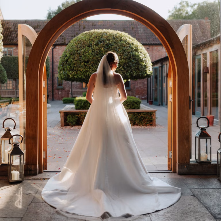 A bride in a white dress and veil stands in a sunlit doorway, facing a courtyard with trimmed trees and brick buildings. Lanterns with candles decorate the entrance.
